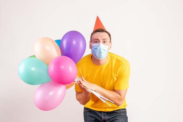 Front view of young man holding colorful balloons in mask on white wall