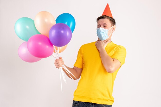 Front view of young man holding colorful balloons in mask on white wall