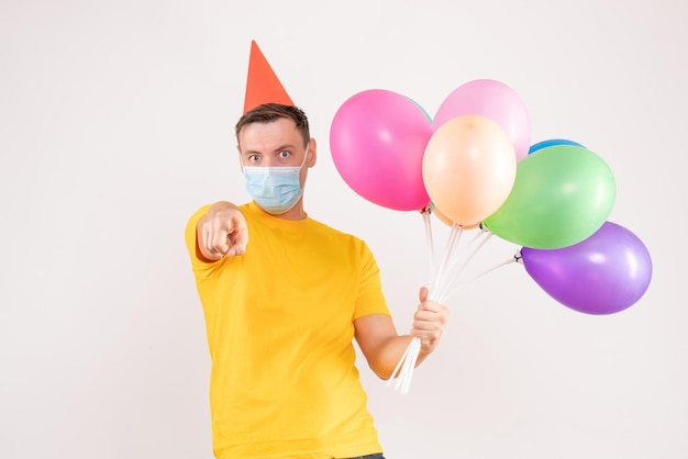 Front view of young man holding colorful balloons in mask on white wall