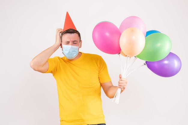 Front view of young man holding colorful balloons in mask on white wall