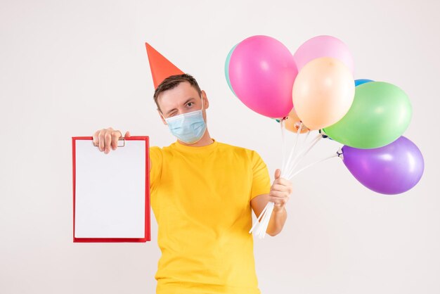 Front view of young man holding colorful balloons in mask on white wall