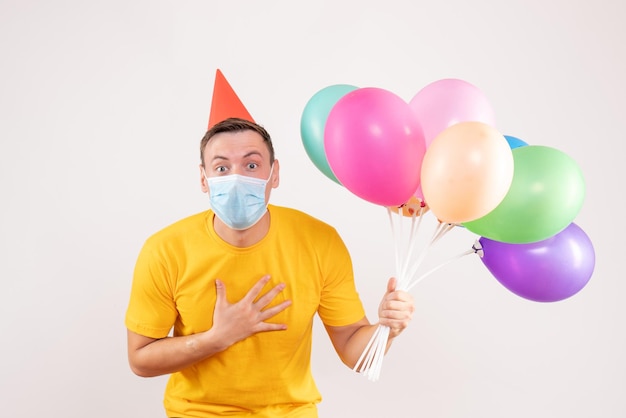 Front view of young man holding colorful balloons in mask on white wall