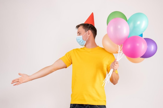 Front view of young man holding colorful balloons in mask on white wall