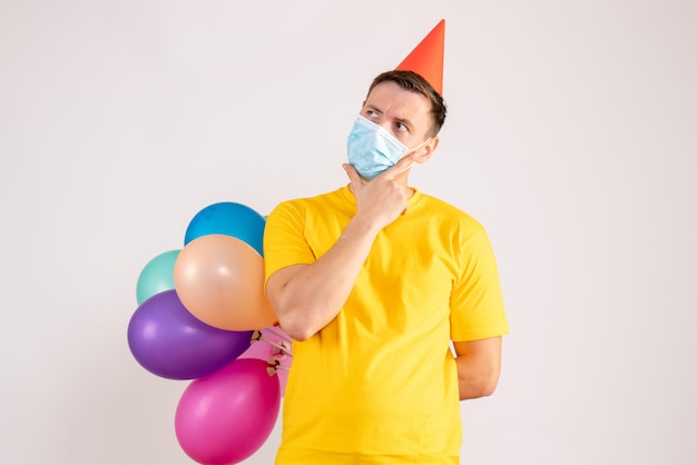 Front view of young man holding colorful balloons in mask on white wall