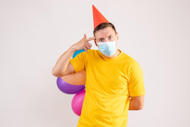 Front view of young man holding colorful balloons in mask on white wall