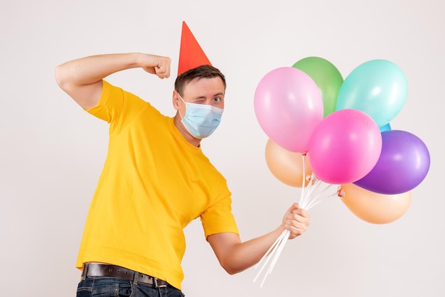 Front view of young man holding colorful balloons in mask on white wall