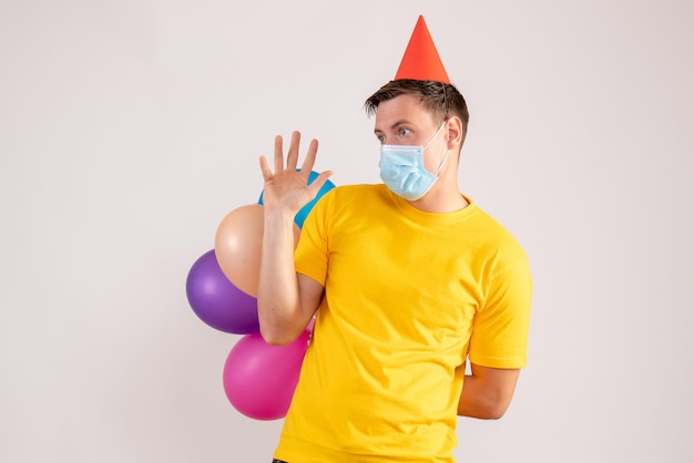 Front view of young man holding colorful balloons in mask on white wall