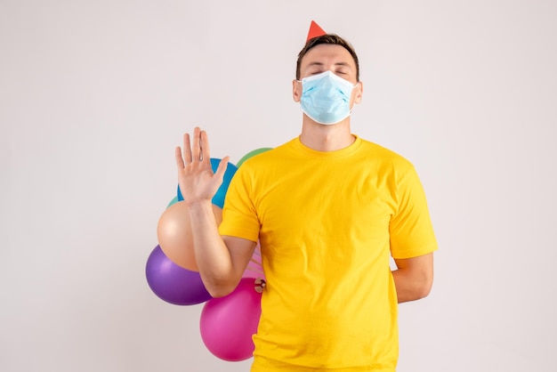 Front view of young man holding colorful balloons in mask on white wall