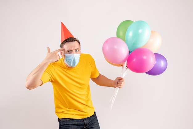 Front view of young man holding colorful balloons in mask on white wall