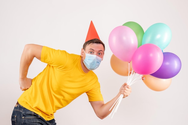 Front view of young man holding colorful balloons in mask on white wall