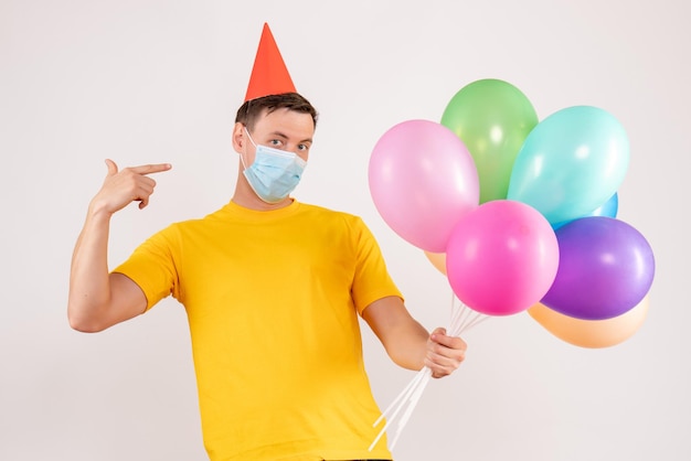 Front view of young man holding colorful balloons in mask on white wall