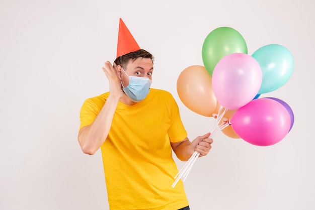 Front view of young man holding colorful balloons in mask on white wall