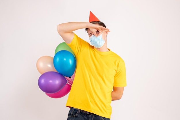 Front view of young man holding colorful balloons in mask on white wall