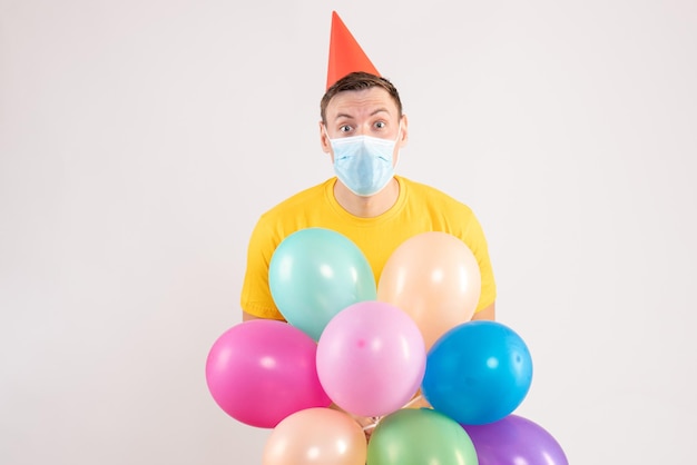 Front view of young man holding colorful balloons in mask on white wall