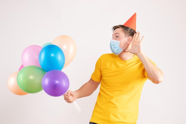 Front view of young man holding colorful balloons in mask on white wall