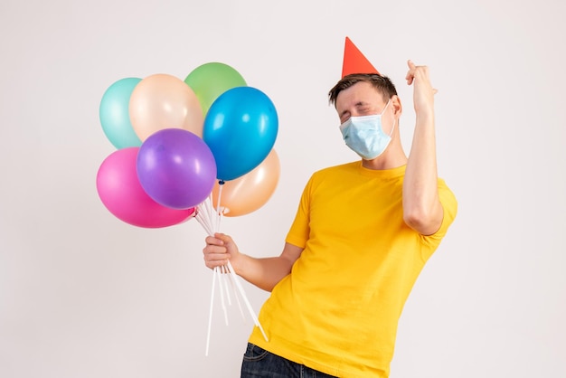 Front view of young man holding colorful balloons in mask on white wall