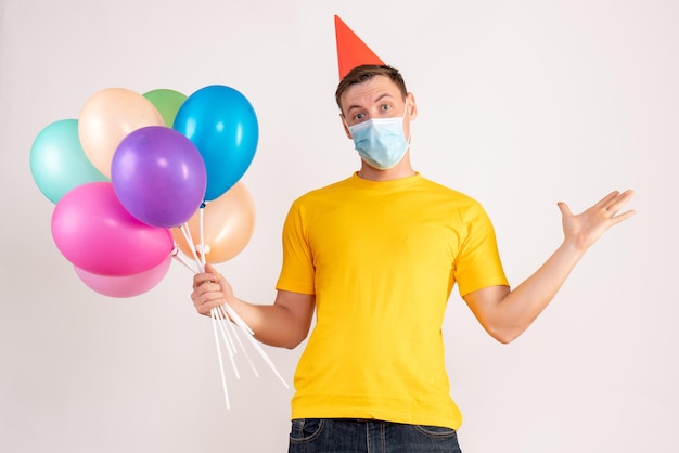 Front view of young man holding colorful balloons in mask on white wall