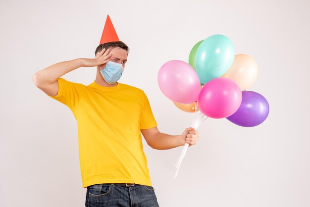 Front view of young man holding colorful balloons in mask on white wall