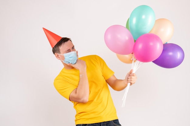 Front view of young man holding colorful balloons in mask on white wall