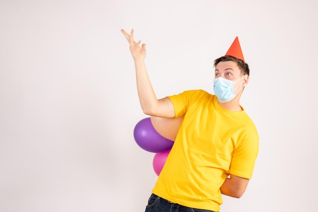 Front view of young man holding colorful balloons in mask on white wall