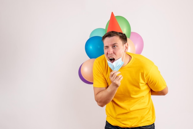 Front view of young man holding colorful balloons in mask on white wall