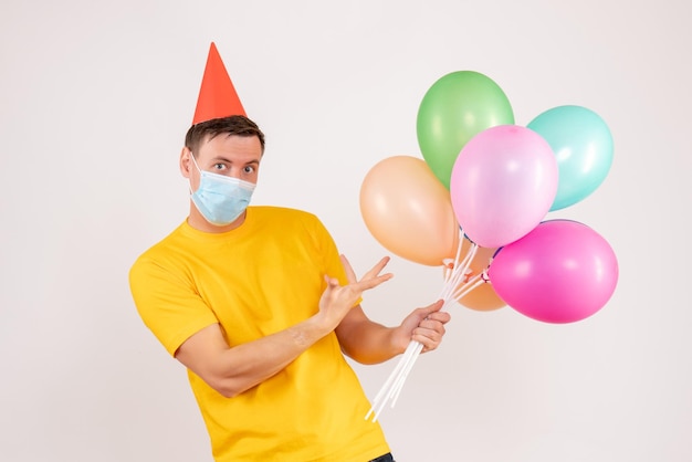 Front view of young man holding colorful balloons in mask on white wall