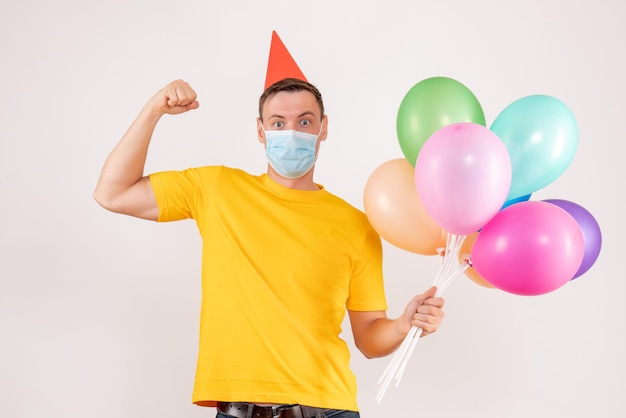 Front view of young man holding colorful balloons in mask on white wall