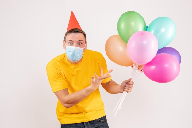 Front view of young man holding colorful balloons in mask on white wall