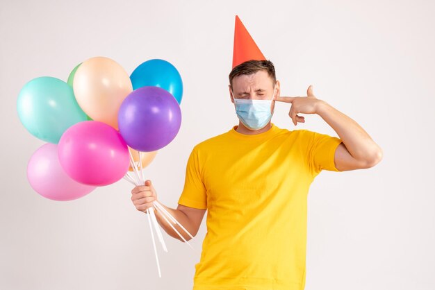 Front view of young man holding colorful balloons in mask on white wall