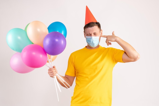 Front view of young man holding colorful balloons in mask on white wall