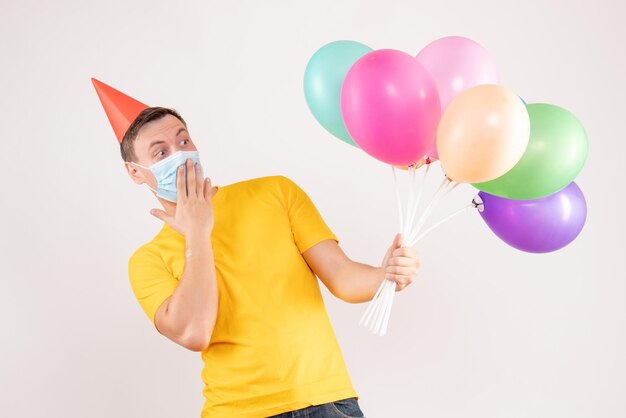 Front view of young man holding colorful balloons in mask on white wall