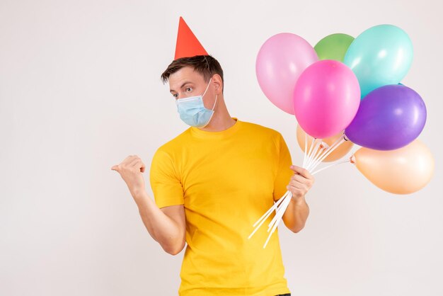 Front view of young man holding colorful balloons in mask on white wall