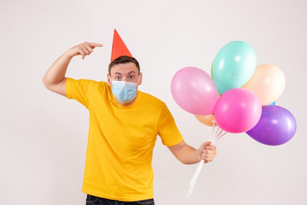 Front view of young man holding colorful balloons in mask on white wall