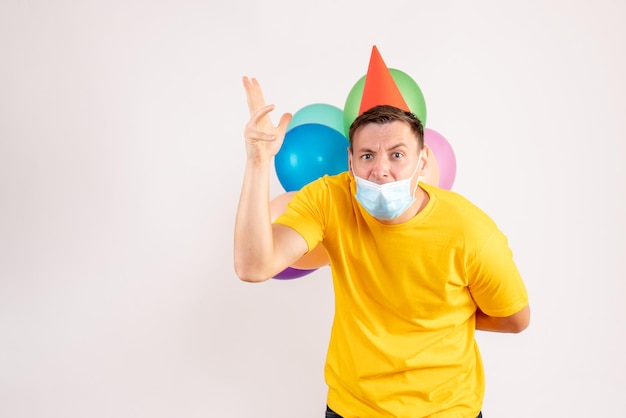 Front view of young man holding colorful balloons in mask on white wall