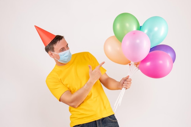 Front view of young man holding colorful balloons in mask on white wall