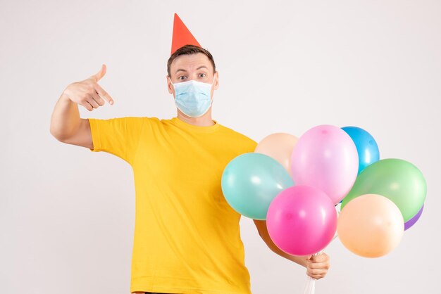 Front view of young man holding colorful balloons in sterile mask on a white wall
