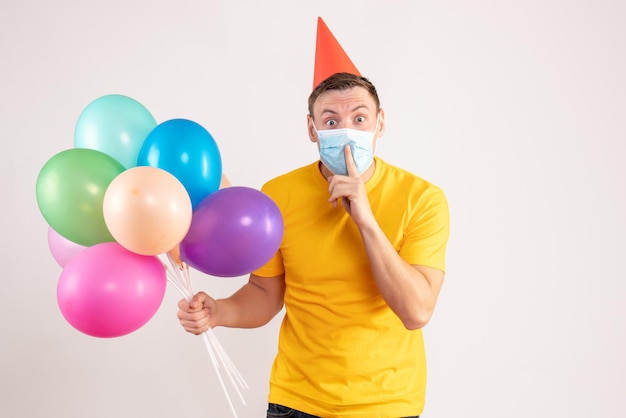 Front view of young man holding colorful balloons in sterile mask on a white wall