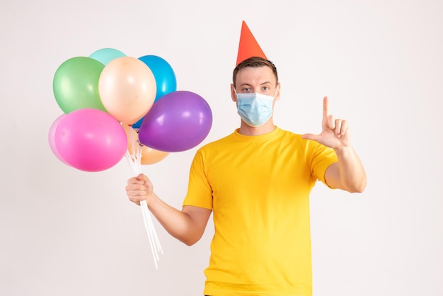 Front view of young man holding colorful balloons in sterile mask on a white wall