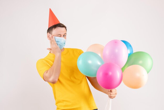 Front view of young man holding colorful balloons in sterile mask on the white wall