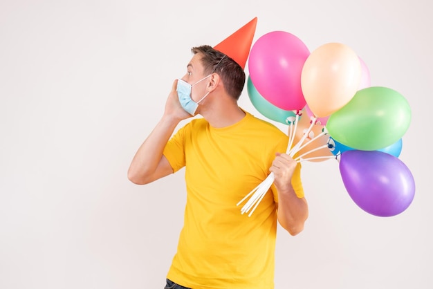 Front view of young man holding colorful balloons in sterile mask on white wall