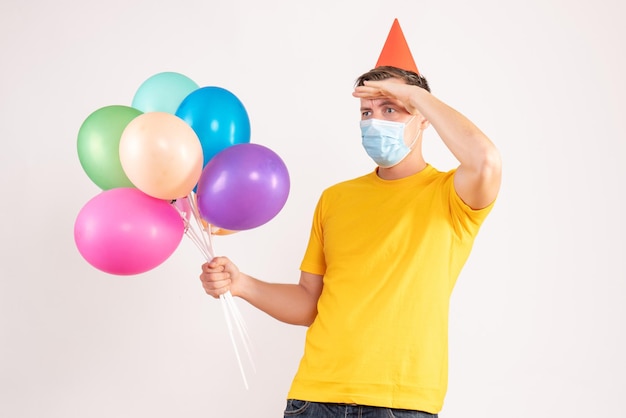 Front view of young man holding colorful balloons in sterile mask on white wall