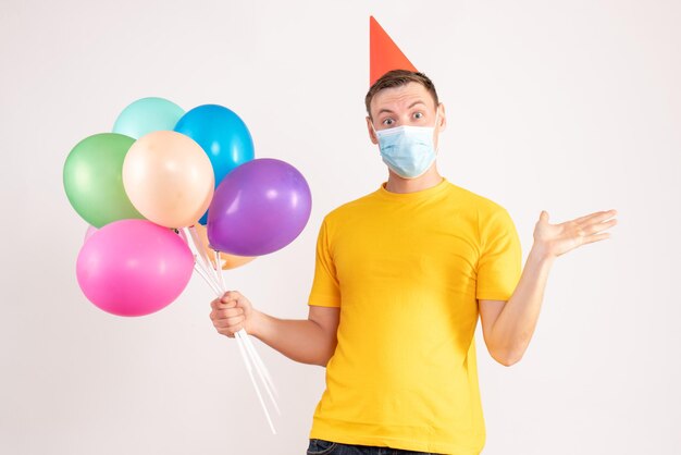 Front view of young man holding colorful balloons in sterile mask on white wall