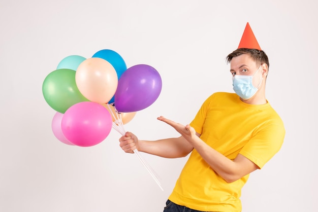 Front view of young man holding colorful balloons in sterile mask on white wall