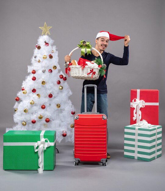 Front view young man holding gift basket and santa hat near xmas tree on grey isolated