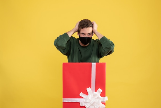 Front view young man holding head standing behind big giftbox on yellow 