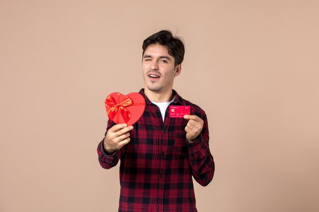 Front view young man holding heart shaped present and bank card on brown wall