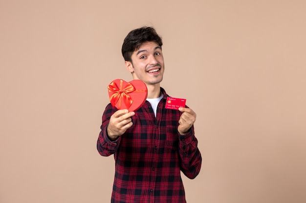 Вид спереди молодой человек, держащий подарок в форме сердца и банковскую карту на коричневой стене