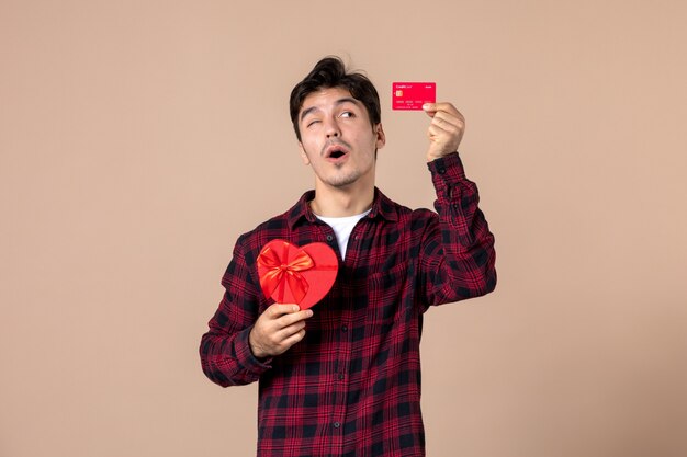 Front view young man holding heart shaped present and bank card on brown wall