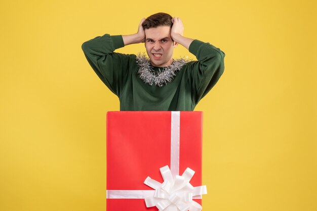 Front view young man holding his head standing behind big giftbox on yellow 