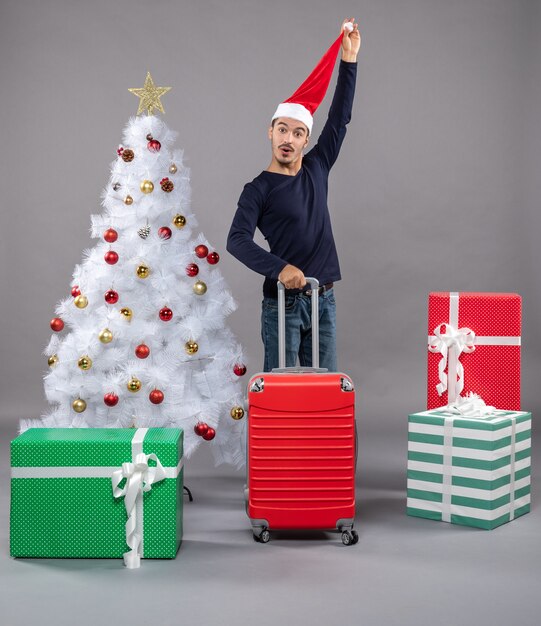 白いクリスマスツリーと灰色の孤立した灰色のカラフルなプレゼントの近くに立っている彼のサンタの帽子を保持している正面図若い男